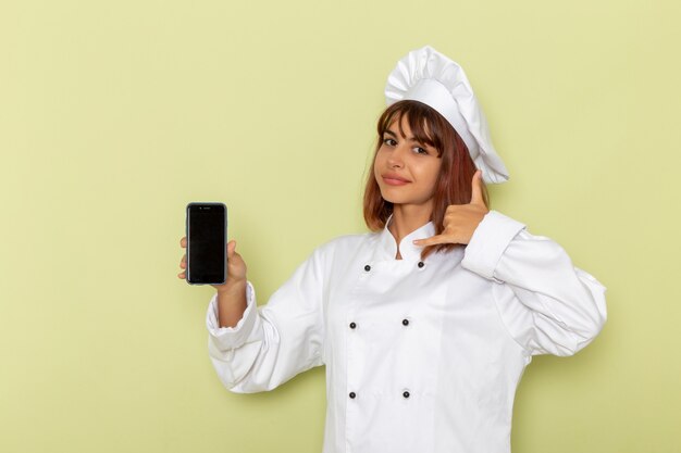 Front view female cook in white cook suit holding her smartphone on green desk