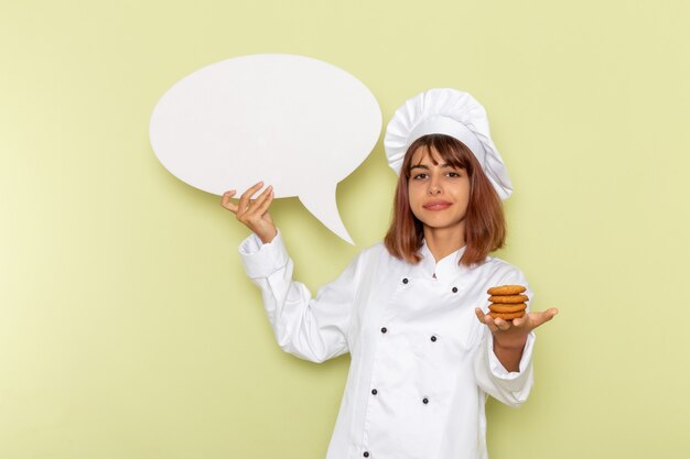 Front view female cook in white cook suit holding little cookies and white sign on green surface