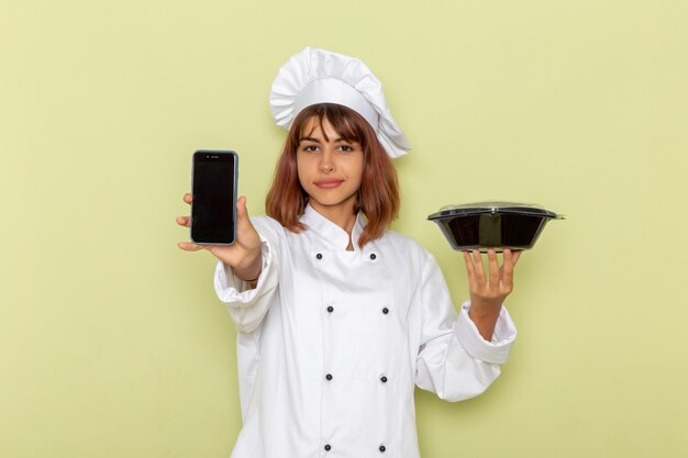 Front view female cook in white cook suit holding phone and black bowl on a green surface