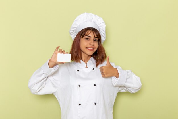 Front view female cook in white cook suit holding white card on light green surface