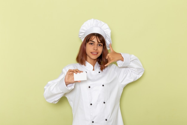 Вид спереди женщина-повар в белом костюме повара держит белую карточку, позирует на зеленой поверхности