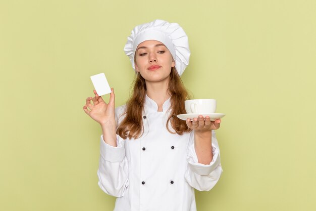 緑の壁に白いプラスチックカードとカップを保持している白いクックスーツの女性料理人の正面図