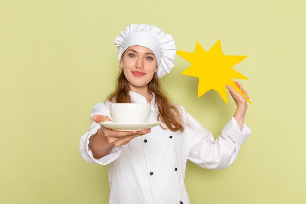緑の壁に黄色の看板とプレートを保持している白いクックスーツの女性料理人の正面図