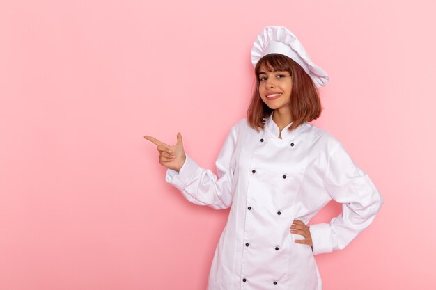 Front view female cook in white cook suit posing and smiling on a pink surface