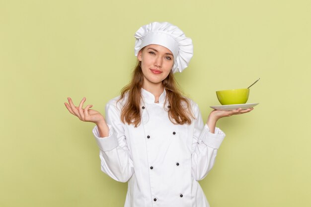 Front view of female cook in white cook suit smiling holding green plate on the green wall