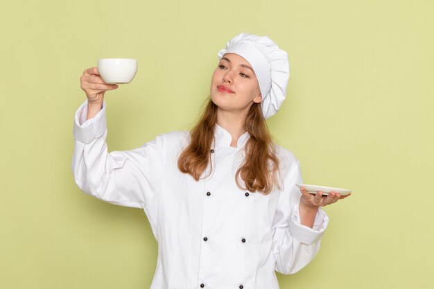 緑の机の上にコーヒーと白いカップを笑顔で保持している白いクックスーツの女性料理人の正面図