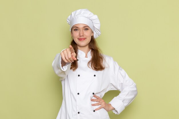 Женщина-повар в белом костюме повара улыбается и показывает на зеленую стену, вид спереди