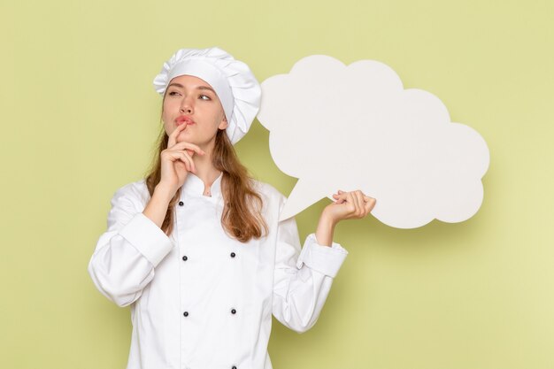 Front view of female cook in white cook suit thinking and holding white sign on green wall