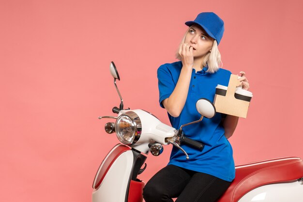Front view female courier on bike with coffee cups thinking on pink 