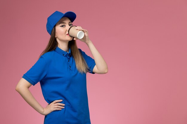 Front view female courier in blue uniform and cape drinking coffee on the pink wall job