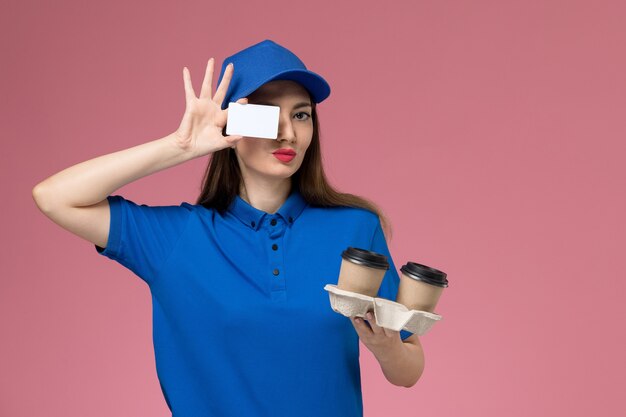 Front view female courier in blue uniform and cape holding brown delivery coffee cups and card on pink wall woman