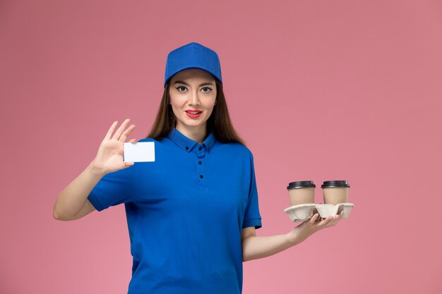 파란색 유니폼과 케이프 핑크 벽에 갈색 배달 커피 컵과 카드를 들고 전면보기 여성 택배