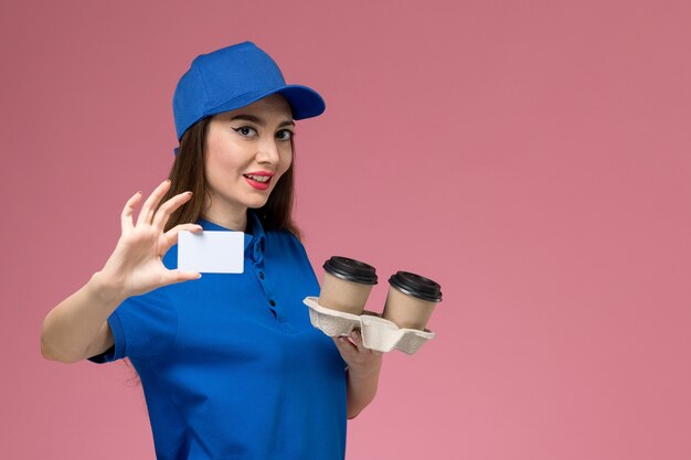 Front view female courier in blue uniform and cape holding brown delivery coffee cups and card with smile on pink wall