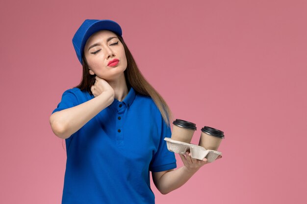 Front view female courier in blue uniform and cape holding brown delivery coffee cups having neckache on pink wall 