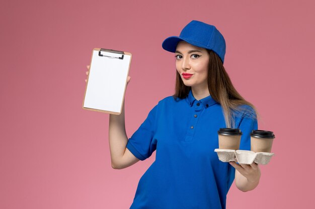 Женщина-курьер в синей форме и плаще, держащая коричневые кофейные чашки доставки и блокнот, улыбается на розовой стене