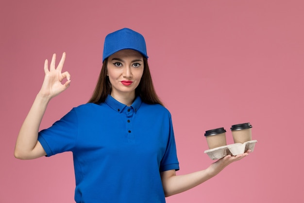 ピンクの机の上に茶色の配達コーヒーカップを保持している青い制服と岬の正面図の女性の宅配便