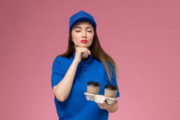 Front view female courier in blue uniform and cape holding brown delivery coffee cups on the pink wall