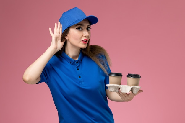 Front view female courier in blue uniform and cape holding brown delivery coffee cups trying to hear on pink wall 