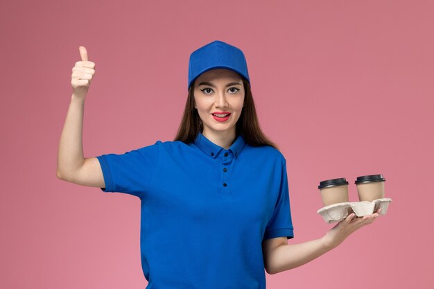 Front view female courier in blue uniform and cape holding brown delivery coffee cups with smile on the light-pink wall