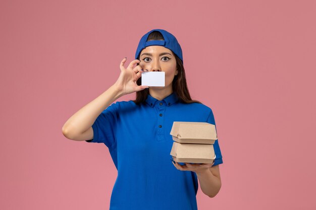 Front view female courier in blue uniform cape holding card and little delivery packages on light pink wall, service employee delivery job