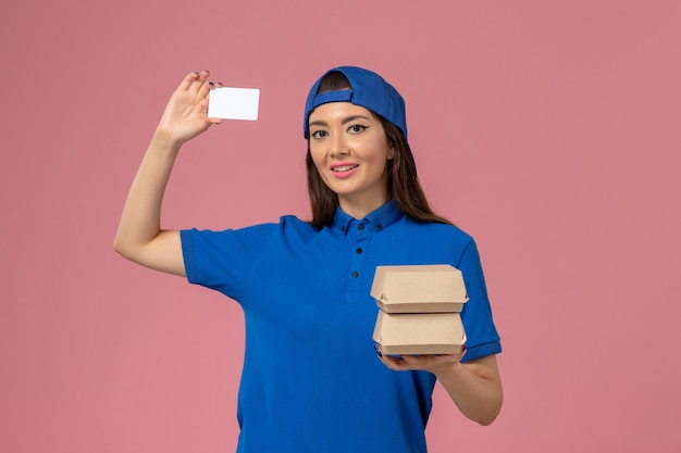 Front view female courier in blue uniform cape holding card and little delivery packages on light pink wall, service employee delivery