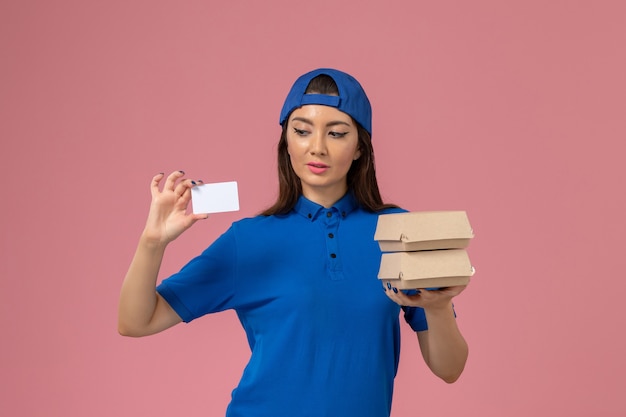 Front view female courier in blue uniform cape holding card and little delivery packages on light pink wall, service job employee delivery