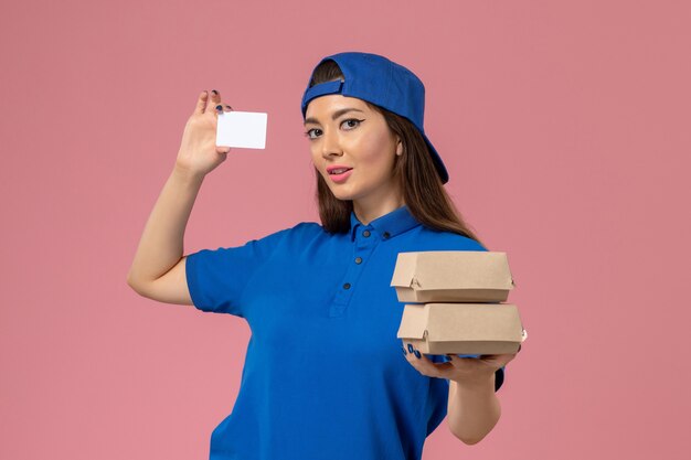 Курьер-женщина, вид спереди в синей форме, держит карточку и маленькие посылки на светло-розовой стене, доставка сотрудника службы доставки