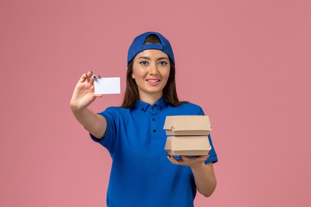 Женщина-курьер в синей форме, держащая карточку и маленькие посылки с доставкой, улыбается на светло-розовой стене, доставка сотрудника службы