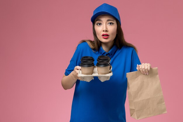 ピンクの壁にコーヒーカップの食品パッケージを保持している青い制服と岬の正面図の女性の宅配便