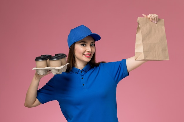 Foto gratuita corriere femminile di vista frontale in uniforme blu e pacchetto dell'alimento delle tazze di caffè della tenuta del capo sulla parete rosa