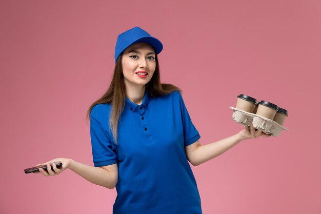 Front view female courier in blue uniform and cape holding coffee cups and smartphone on pink wall 