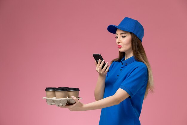 Front view female courier in blue uniform and cape holding coffee cups taking a photo on pink wall 