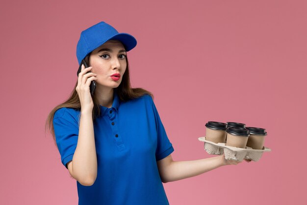 Женщина-курьер в синей форме и плаще, держащая кофейные чашки, разговаривает по телефону на розовой стене, вид спереди