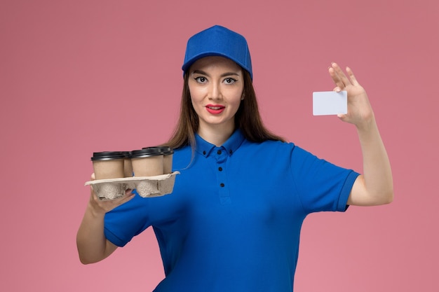 Front view female courier in blue uniform and cape holding coffee cups and white card on pink wall 