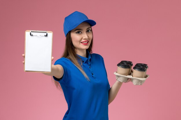 Front view female courier in blue uniform and cape holding coffee cups with notepad on pink wall  woman