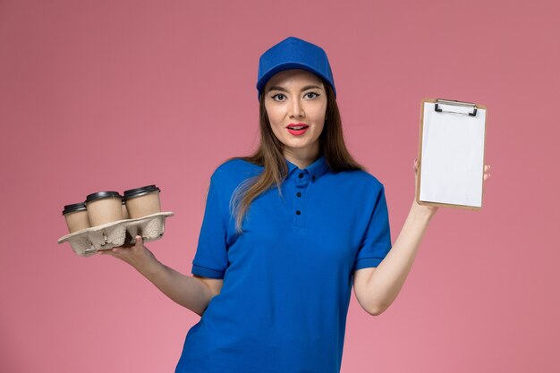 ピンクの壁の仕事の女性の女性にメモ帳とコーヒーカップを保持している青い制服と岬の正面図の女性の宅配便