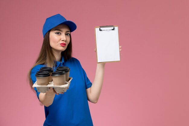 Front view female courier in blue uniform and cape holding coffee cups with notepad on pink wall  work woman