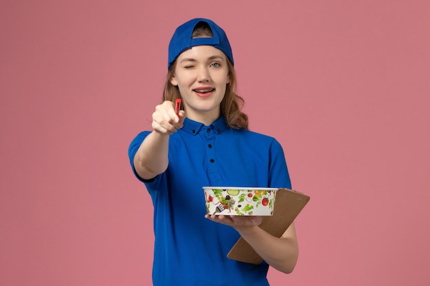 Corriere femminile di vista frontale in capo uniforme blu che tiene la ciotola di consegna e la penna del blocco note sulla parete rosa-chiaro, impiegato del lavoro di consegna di servizio