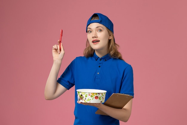 淡いピンクの壁に配達ボウルとメモ帳のペンを保持している青い制服ケープの正面図女性宅配便、サービス配達労働者の従業員