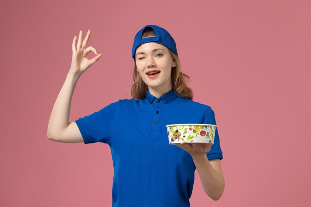 Front view female courier in blue uniform cape holding delivery bowl winking on pink wall, worker service delivery employee