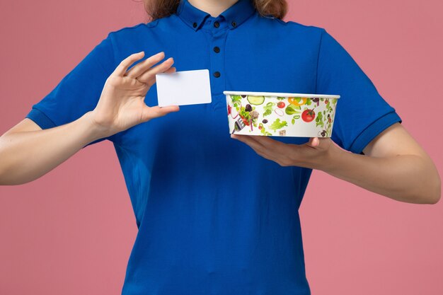 Front view female courier in blue uniform cape holding delivery bowl with card on light pink wall, service delivery employee job
