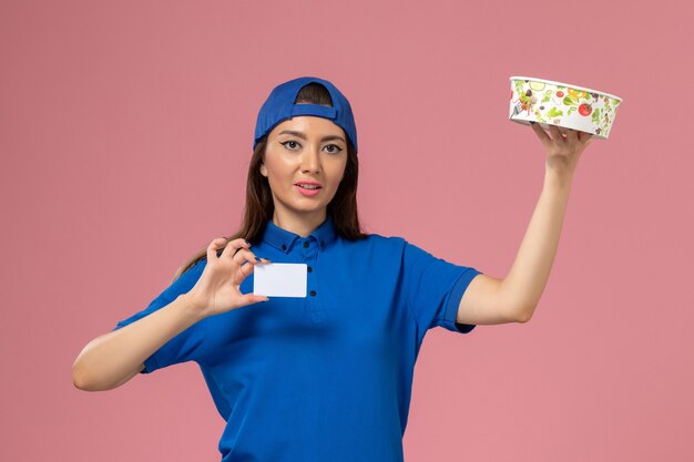 Front view female courier in blue uniform cape holding delivery bowl with card on light-pink wall, service employee delivery work