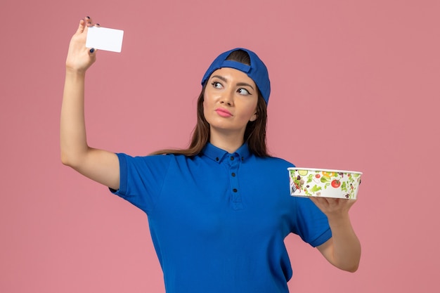 Женщина-курьер в синей форме, держащая миску с карточкой на светло-розовой стене, работник службы доставки