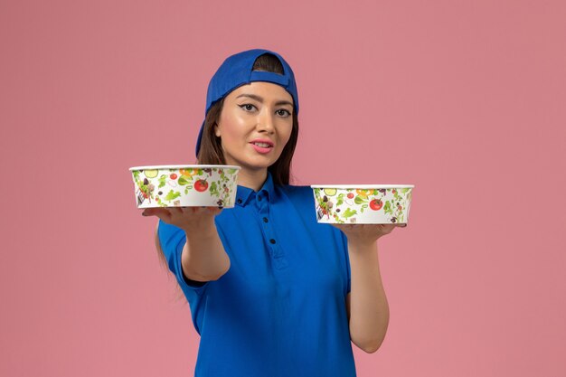 Front view female courier in blue uniform cape holding delivery bowls on light pink wall, service employee delivery job