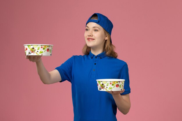 Front view female courier in blue uniform cape holding delivery bowls on pink wall, service delivery job employee