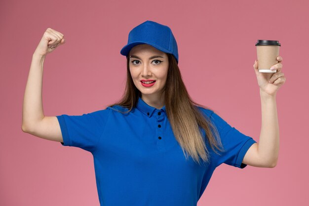 Front view female courier in blue uniform and cape holding delivery coffee cup and flexing on the pink wall job