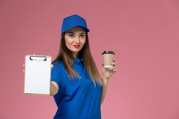 Front view female courier in blue uniform and cape holding delivery coffee cup and notepad on the light-pink desk 