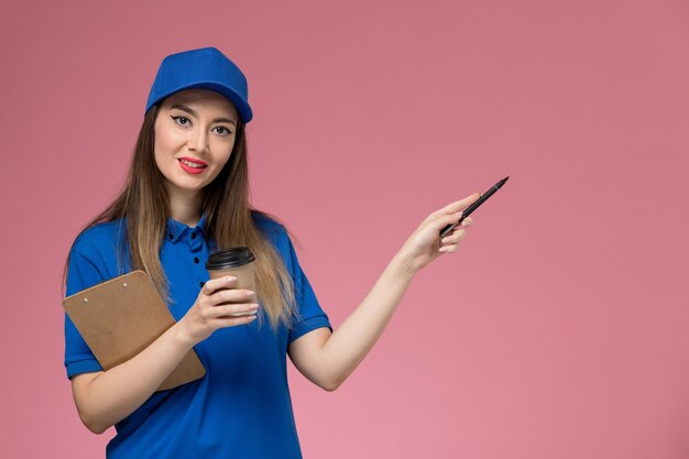 Front view female courier in blue uniform and cape holding delivery coffee cup notepad and pen on pink wall