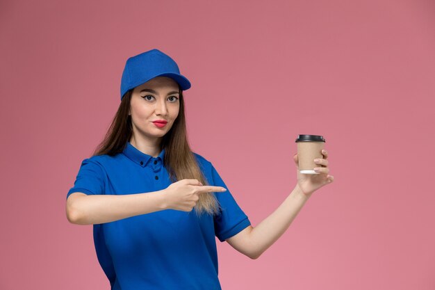 Front view female courier in blue uniform and cape holding delivery coffee cup on the pink wall girl woman worker work