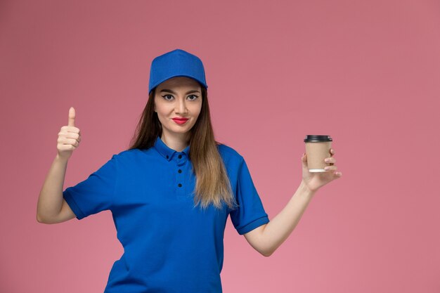 Front view female courier in blue uniform and cape holding delivery coffee cup on pink wall job woman